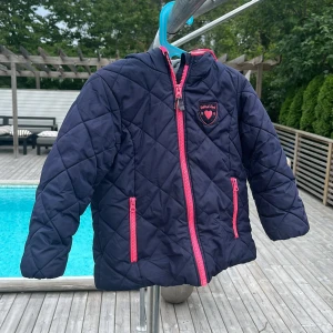 Quiltad blå jacka från OshKosh, stl 6 - Mörkblå quiltad jacka från OshKosh B'gosh med rosa dragkedjor och detaljer. Jackan har huva, två fickor med dragkedja och ett mjukt grått foder. 