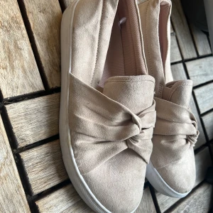Beige slip-on sneakers med rosett - Snygga slip-on sneakers i beige mocka-look med bred rosettdetalj framtill. Skorna har vit platt sula och rund tå, vilket ger en clean och trendig vibe. Perfekta för dig som gillar stilrena men ändå unika skor.