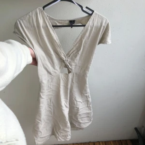 Beige kortklänning med knyt i ryggen - Superfin beige kortklänning från i bomull med v-ringning och öppen rygg som man knyter. Klänningen har korta ärmar och lätt tunt material. den är M men passar S oxå