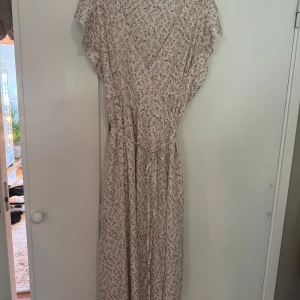 Ljusblommig långklänning med volangärm - Superfin långklänning i ljusbeige med små rosa blommor och gröna blad. Klänningen har omlottliv, volangärmar och knytband i midjan för en snygg siluett. Perfekt för sommarens alla tillfällen! 