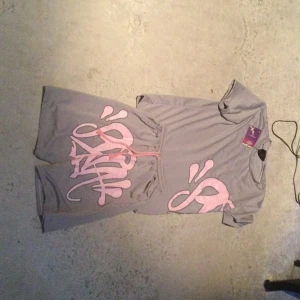 Syna World - Grå shorts från Syna World med stort rosa grafiskt tryck framtill och matchande snörning i midjan. De är tillverkade i ett mjukt syntetmaterial och har en loose passform som ger en avslappnad vibe. Perfekta för chill dagar eller när du vill sticka ut lite extra.