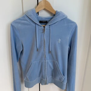 Ljusblå velour hoodie Juicy Couture - Supermjuk ljusblå hoodie från Juicy Couture i velour med dragkedja och huva. Klassisk broderad JC-logga på bröstet, snörning i huvan och känguruficka framtill. 
