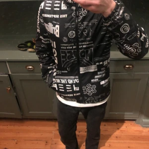 Kenzo vindjacka - Svart kenzo vindjacka med huva och hel dragkedja framtill. Jackan har ett unikt grafiskt tryck i vitt med olika texter och symboler över hela ytan. Tillverkad i lätt polyester, perfekt för dig som vill sticka ut med en cool streetstyle-look.