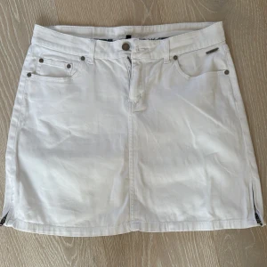 Vit jeanskjol från Sebago - Vit jeanskjol från Sebago med klassisk femficksdesign, bälteshällor och dragkedja fram. Kjolen är kort och har små slitsar i sidorna. Perfekt för dig som gillar en clean och fräsch look.