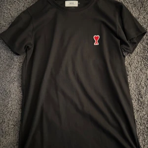 Svart t-shirt från Ami Paris med hjärtdetalj - Svart t-shirt från Ami Paris med klassisk passform och korta ärmar. På bröstet sitter Ami:s ikoniska röda hjärta med A-bokstav. Tillverkad i mjuk bomull och har broderad AMI-logga i nacken. Perfekt för en clean och stilren look.