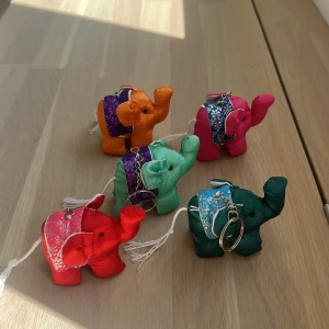 Färgglada elefant-nyckelringar i tyg - Fem små elefantformade nyckelringar i tyg, i färgerna röd, grön, orange, rosa och mörkgrön. Varje elefant har ett dekorativt tygsadel i glittriga färger och mönster samt en vit snodd. Perfekt som accessoar till väskan eller nyckelknippan. EN FÖR 39❤️❤️