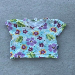 Blommig kortärmad topp från NQT - Supergullig kortärmad topp från NQT med färgglatt blommönster i lila, gult och rött på ljusblå botten. Toppen har rund hals och är tillverkad i mjuk bomull, perfekt för sommarens alla äventyr.