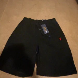Svarta shorts Polo Ralph Lauren S - Svarta shorts från Polo Ralph Lauren med klassisk röd broderad logga på benet. Mjuk bomullskänsla, elastisk midja med snörning för skön passform. Perfekta för chill dagar eller när du vill ha en clean och enkel stil.