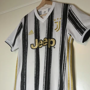 Juventus Adidas fotbollströja svartvit - Juventus matchtröja från Adidas med svartvita ränder och guldiga detaljer. Jeep-logga på bröstet, klubbmärke med tre stjärnor och Adidas-logga i guld. Kortärmad modell i lätt och ventilerande material, perfekt för fotbollsplanen.