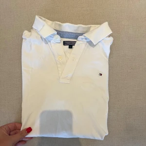 Vit pikétröja från Tommy Hilfiger - Klassisk vit pikétröja från Tommy Hilfiger med liten broderad logga på bröstet. Tröjan har krage, korta ärmar och knäppning med två knappar framtill. Tillverkad i mjuk bomull som känns skön mot huden. Perfekt för en clean och stilren look. Storlek 176 motsvarar m/s