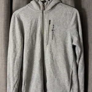 Grå hoodie från Sail Racing med dragkedja - Snygg ljusgrå hoodie från Sail Racing med hel dragkedja och broderad logga på bröstet. Tröjan har huva, lång ärm och en bröstficka med dragkedja. Mjuk insida i fleece som gör den extra skön. 
