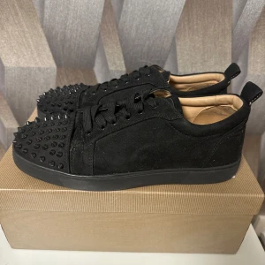 Louboutin skor - Svarta sneakers i mocka från Christian Louboutin med coola svarta nitar på tån och klassisk röd sula. Skorna har snörning och en platt sula, perfekt för dig som vill sticka ut med en edgy vibe. Insidan är beige och materialet känns lyxigt.