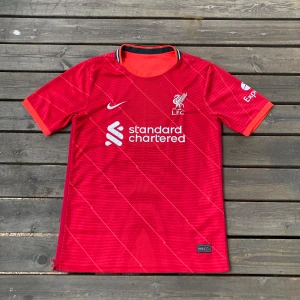 Liverpool FC röd matchtröja Nike - Liverpool FCs officiella matchtröja från Nike i rött med subtila diagonala linjer och vita logotyper. Kortärmad modell med svart och vitt band i kragen. Tillverkad i lätt och ventilerande Dri-FIT-material, perfekt för fotboll eller supporterliv.