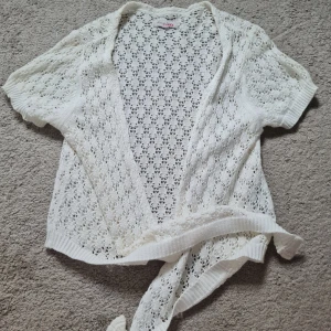 Vit stickad kofta med knyt från Claina - Supersöt vit stickad kofta från Claina med korta ärmar och knytband framtill. Koftan har ett genomgående hålmönster och ribbade kanter. Perfekt att slänga över ett linne för en chill och somrig look.