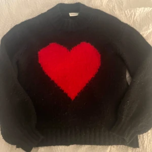 Svart stickad tröja med rött hjärta Kappahl - Mysig svart stickad tröja från Kappahl med ett stort rött hjärta på bröstet. Tröjan har rund halsringning, ribbade muddar och lång ärm. Perfekt för dig som vill ha en statement-piece i garderoben. Jättefin som passar till mycket 