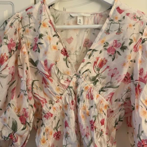Blommig kortklänning från H&M - Supersöt kortklänning med blommigt mönster i rosa, gul och grönt på ljus botten. Klänningen har v-ringning, puffiga trekvartsärmar och smockad midja som ger en snygg passform. Perfekt för vår och sommar!