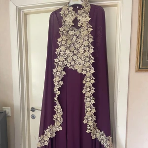 Lång lila klänning med spetsdetaljer - Elegant långklänning i djup lila med tunna axelband och matchande cape. Klänningen har vackra guldiga spetsapplikationer med blommönster och pärlor över byst och cape. Perfekt för bal eller fest, ger en lyxig och unik look.