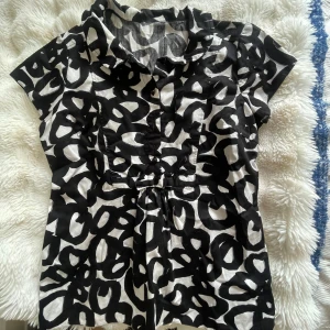Svartvit blus med grafiskt mönster - Snygg svartvit blus med coolt grafiskt mönster och korta ärmar. Blusen har krage, knappar framtill och lätt insvängd midja för en schysst passform. Perfekt för dig som gillar att sticka ut med din stil. 