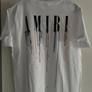 Vit AMIRI t-shirt med färgstänk - Vit t-shirt från AMIRI med tryck av loggan och färgstänk i blått, rött, orange och svart som rinner ner över både fram- och baksida. Klassisk passform och rund hals. Perfekt för dig som gillar streetstyle och unika detaljer.