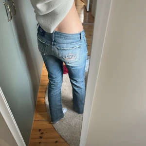 Blåa lågmidjade jeans från Rock & Republic - Snygga blå jeans från Victoria Beckham med låg midja och bootcut. Jeansen har coola detaljer som broderad krona på bakfickorna och en utbytt metallknapp framtill då original knappen åkte av. Förra ägaren har sytt ihop ett stort hål i bak och sytt in midjan med ca 6cm sammanlagt, inget man tänker på när dom är på(se bild 5) midja:37cm innerbensmåtten:77cm