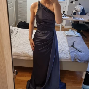 Mörkblå långklänning med slits - Snygg mörkblå långklänning i satinliknande material med enaxlad design och pärldetaljer längs axel och midja. Klänningen har en hög slits framtill och markerad midja som ger en elegant siluett. Perfekt för bal eller festliga tillfällen. Den var köpt för cirka 3 000kr och endast använd en gång.