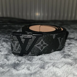 Louis Vuitton svart skärp med monogram - Snyggt svart Louis Vuitton-bälte med klassiskt LV-monogram i grått och stor LV-logga som spänne. Bältet är gjort i präglat skinn och har en stilren, modern look som sticker ut. Perfekt accessoar för att lyfta din outfit med exklusiv känsla.