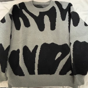Grå stickad tröja - Säljer en grå stickad tröja med unikt svart abstrakt mönster. Tröjan har rund halsringning, ribbade muddar och är långärmad. Tröjan är knappt använd och i storlek L men liten i storleken så passar mer M/S