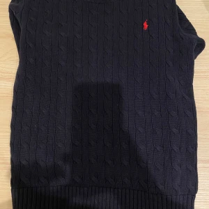 Mörkblå stickad tröja Polo Ralph Lauren - Kabelstickad mörkblå tröja från Polo Ralph Lauren med röd broderad logga på bröstet. Tröjan har rund halsringning och ribbade muddar. Väldigt bra för hösten.