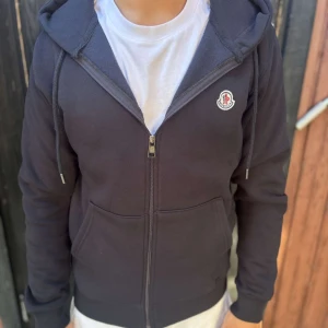 Svart hoodie från Moncler med dragkedja - Svart hoodie från Moncler med klassisk logga på bröstet, hel dragkedja och två stora fickor framtill. Tröjan har huva med snörning och är tillverkad i mjuk bomull. Perfekt för en chill och stilren look.