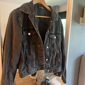 Svart jeansjacka från Divided, strl 36 - Snygg svart jeansjacka från Divided i klassisk modell med silverfärgade knappar och två bröstfickor. Jackan har en rak passform och är tillverkad i denim. Perfekt att slänga över en hoodie eller t-shirt för en chill look.