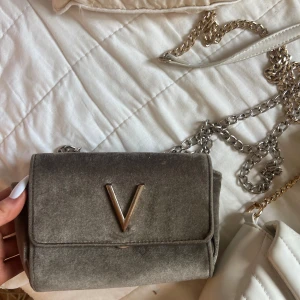 Grå axelväska i sammet från Valentino - Snygg grå axelväska från Valentino i mjuk sammet med silverfärgad kedjerem och ett stort V i metall framtill. Perfekt storlek för det viktigaste och har ett innerfack med dragkedja. Stilren och trendig accessoar som lyfter din outfit.