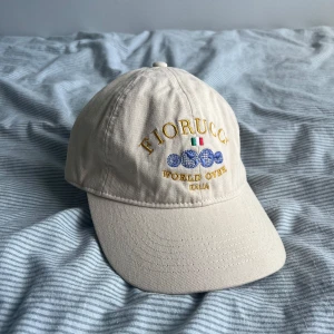 Creme-vit keps från Fiorucci - Snygg keps från Fiorucci med broderad logga och text i gult framtill samt italiensk flagga och blå detaljer. Justerbar rem baktill med metallspänne där det står Fiorucci. Tillverkad i 100% bomull. Knappt använd och i mycket bra skick. Tveka inte på att höra av dig vid frågor eller funderingar!