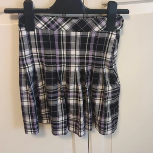 Lila och svart rutig kjol från Lindex - Supersöt tkjol från Lindex i rutigt mönster med lila, svart och vitt. Kjolen har veckad design och stängs med knapp och dragkedja i sidan.💞