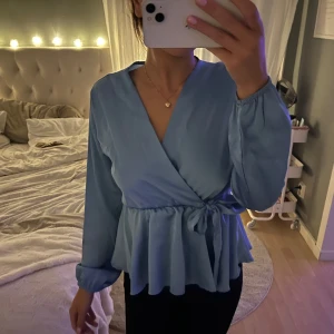 Ljusblå omlottblus med knyt - Supersöt ljusblå omlottblus med v-ringning och knyt i midjan. Blusen har långa ärmar med resår vid handlederna och volang nedtill som ger en flowy känsla. Den har dessvärre några fläckar men det är verkligen inte som egentligen syns🩷