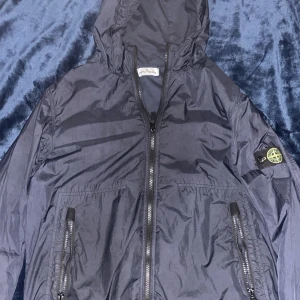 Svart windbreaker från Stone Island - Snygg svart vindjacka från Stone Island med huva och dragkedja framtill. Jackan har två fickor med dragkedja och den klassiska loggan på vänster ärm. Perfekt för dig som vill ha en stilren och sportig look.