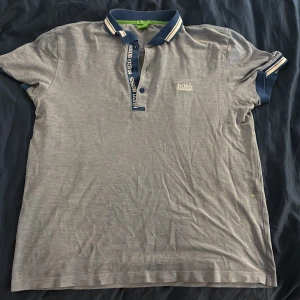 Grå pikétröja från Hugo Boss - Snygg grå pikétröja från Hugo Boss med blå och vita detaljer på krage och ärmslut. Klassisk pikéknäppning med logga längs knappraden och broderad Boss-logga på bröstet. Perfekt för en clean och sportig stil.