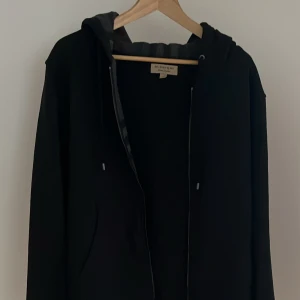Svart Burberry Zip up - Svart tröja från Burberry med dragkedja och huva. Insidan av huvan har klassiskt rutigt Burberry-mönster i grått och rött. Tröjan  har snörning i huvan och två fickor framtill. Perfekt för dig som vill ha en stilren och ikonisk look.