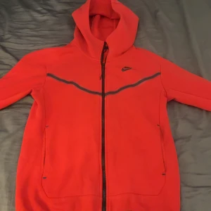 Röd Nike hoodiejacka XS - Nike hoodiejacka i rött med svart logga på bröstet och hel dragkedja framtill. Jackan har huva, långa ärmar och svarta detaljer längs dragkedjan och fickorna. Perfekt för dig som gillar sportig och avslappnad stil. Lite syd på ärmen.