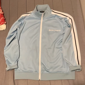 Palm Angels ljusblå track jacket - Palm Angels ljusblå track jacket med vita och svarta ränder längs ärmarna. Jackan har hel dragkedja framtill, hög krage och logga på bröstet. Tillverkad i polyester för en sportig vibe och skön passform. Perfekt för dig som gillar streetwear och snygga detaljer.