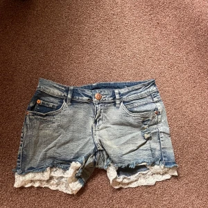 Ljusblå spets shorts  - Snygga blå jeansshorts med slitna detaljer och vit spetskant längst ner. Klassisk femficksmodell med knapp och dragkedja. Perfekta för sommaren och ger en cool, feminin vibe till din outfit.