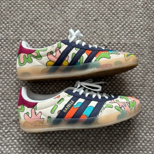 Adidas Gazelle x Sean Wotherspoon sneakers - Färgglada Adidas Gazelle sneakers i samarbete med Sean Wotherspoon. Skorna har ett unikt, broderat mönster i flera färger på manchestertyg, mörkblå ränder och vit snörning. Sula i gummi och detaljer i vinrött på hälen. Perfekta för dig som vill sticka ut. Säljer dessa pga att dom bara har använts en gång. 