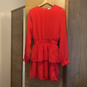 Röd volangklänning från Kappahl - Superfin röd kortklänning från Kappahl med långa ärmar, markerad midja med smock och volangdetaljer i kjolen. Klänningen har rund halsringning och stängs med knapp i nacken. Perfekt för dig som vill sticka ut med färg!