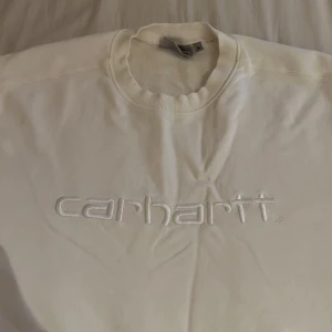Vit Carhartt sweatshirt med broderad logga - Säljer en stilren vit sweatshirt från Carhartt med broderad logga framtill. Tröjan har rund halsringning, långa ärmar och är gjord i mjuk bomull. Perfekt för dig som gillar clean och enkel streetwear.