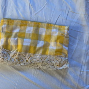 Gul och vit rutig kuvertväska med spets - Söt kuvertväska i gul och vit rutig bomull med dragkedja upptill. Väskan har en dekorativ spetskant i vitt längs nederkanten som ger en vintagekänsla. Perfekt för att förvara småsaker eller smink i väskan.