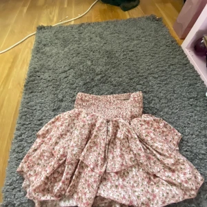 Rosa blommig volangkjol - Supersöt kort kjol i rosa med små blommor och volanger. Kjolen har en smockad midja som sitter bekvämt och ger en fin passform. Lätt och luftig, passar perfekt till varma dagar. Pris kan diskuteras.