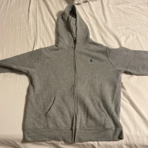 Grå hoodie från Polo Ralph Lauren - Ge mig ett bud, jag är inte fast vid pris:)Klassisk grå hoodie från Polo Ralph Lauren med dragkedja och huva. Tröjan har två fickor framtill och den ikoniska lilla broderade loggan på bröstet. Perfekt för chill dagar och enkel att matcha med jeans eller joggers. Säljer pågrund av att den är för liten, jag är 176 och den är liten på mig. Nypris 1200, Pris går att diskutera!