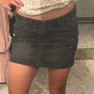 Svart jeanskjol med slitningar - Snygg svart jeanskjol i kort modell med slitna detaljer framtill och bakfickor. Kjolen har klassisk femficksdesign och en cool, lite tvättad look. Perfekt för dig som gillar en edgy och avslappnad stil.