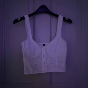 Vit broderad korsett från Divided - Superfin vit korsett-topp från Divided i storlek 38. Toppen är broderad med små hålmönster och har breda axelband samt markerade kupor. Perfekt till sommaren och ger en fräsch och trendig look med sin croppade passform.