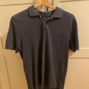 Mörkblå pikétröja från Boomerang - Snygg mörkblå pikétröja från Boomerang med klassisk krage och två knappar framtill. Tröjan är i bomull och har korta ärmar, perfekt för en clean och stilren look. Liten broderad logga på bröstet ger en diskret touch.