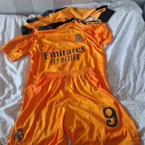 Real Madrid Mbappé fotbollströja orange - Säljer en orange Real Madrid fotbollströja med Mbappé och nummer 9 på ryggen. Setet inkluderar matchande shorts och strumpor. Tröjan har korta ärmar framtill och långa ärmar baktill, med svarta detaljer och klubbmärke på bröstet. Materialet är lätt och andas, perfekt för fotboll.